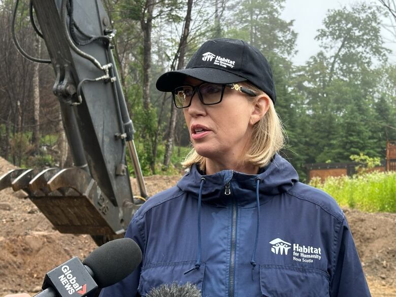 A woman in a ble raincoat wearing a Habitat for Humanity cap speaks into a microphone