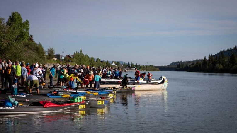 A crowd of people and canoes and kayaks are seen gathered on a river bank.