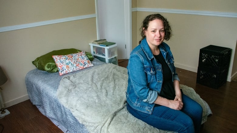 A woman in a blue jean jacket sits on her twin bed with her hands folded. Black milk crates are seen in the background. 