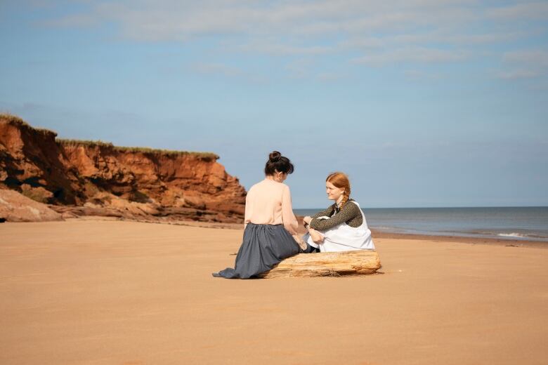 Two women in old-fashioned clothing sit together on the beach. 