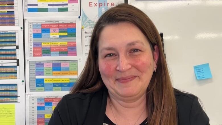 A smiling woman sits behind her desk with colour-coded schedules on the wall behind her.