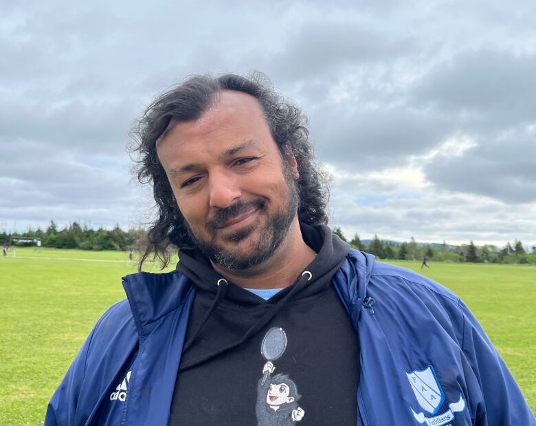 A man smiles. In the background, people play cricket on a field.