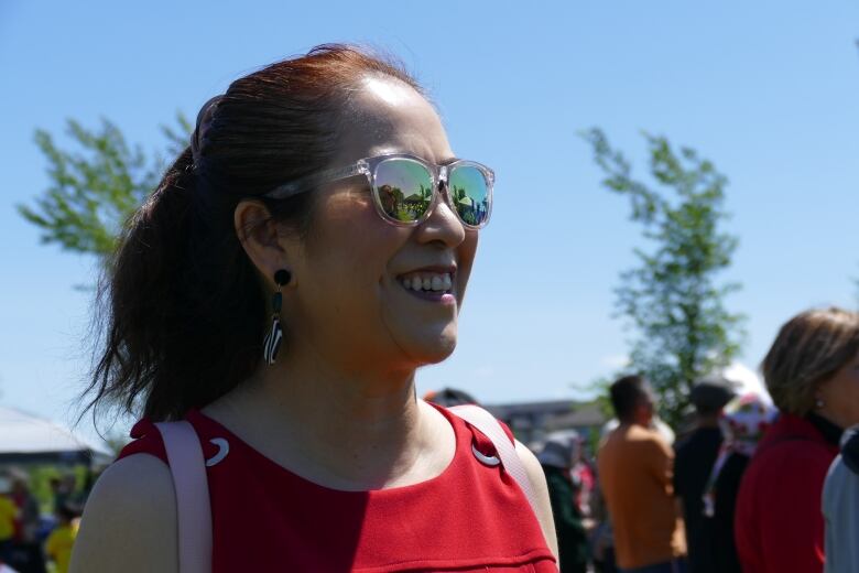 A woman wearing a red shirt and sunglasses smiles while looking away from the camera. 