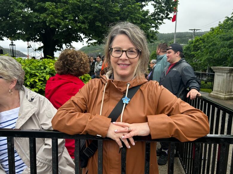 Woman in brown rain coat with blue flower pinned, leaning against rail.