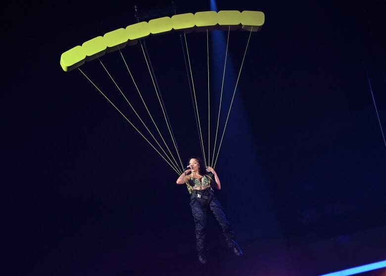 A woman holding a microphone is suspended in the air, attached to a prop parachute. 