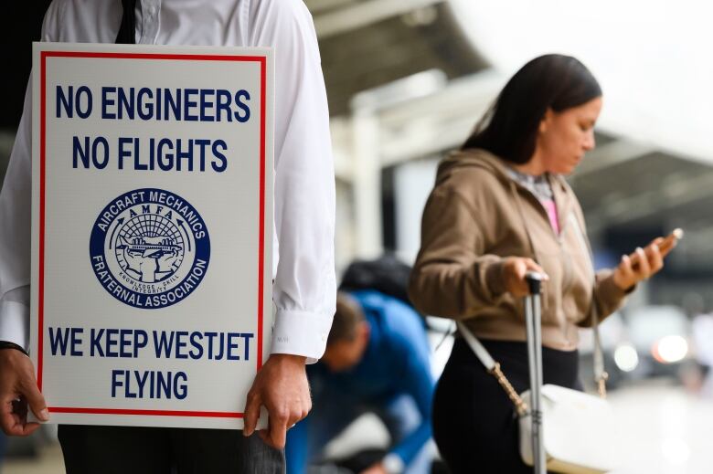 A striking worker's sign is seen with a woman checking her phone in the background.