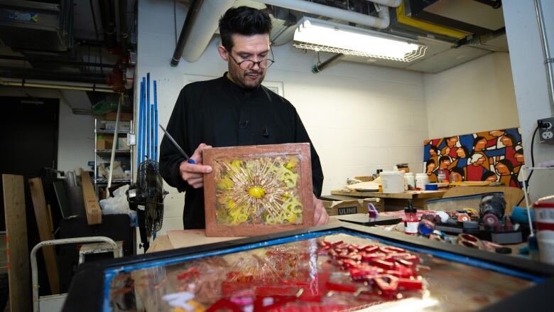 A man looks at glass pieces while making artwork, with other art equipment around him.