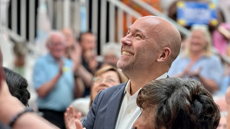 A man wearing a navy suit and white collared shirt looks upward and smiles broadly in a crowd of people.