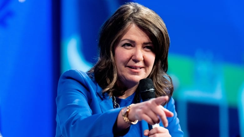 A woman wearing a blue blazer points at someone in an audience.