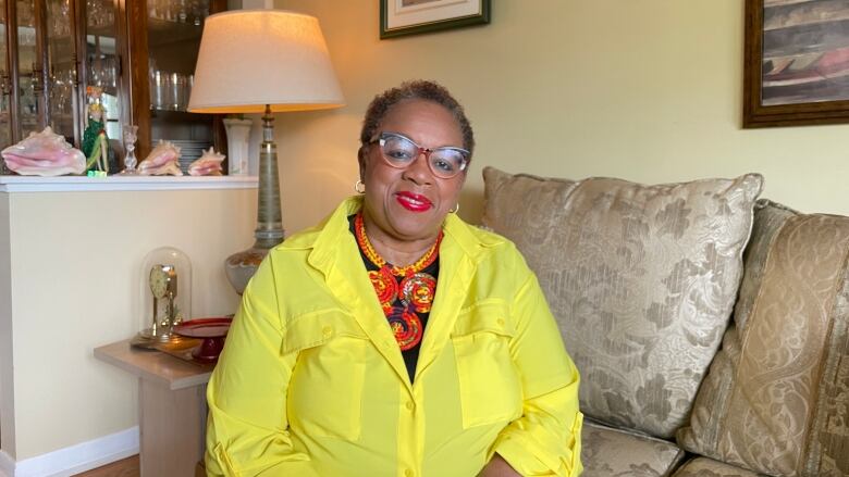 A woman sits on a couch in her house wearing a bright yellow shirt and red lipstick. 