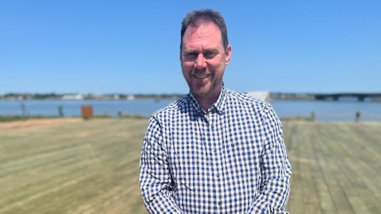 Man with beard, wearing a blue checked shirt, stands on wooden platform overlooking a river. 