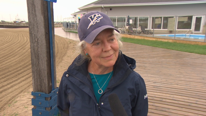 A woman wearing a navy raincoat speaks into a microphone near a beach. 