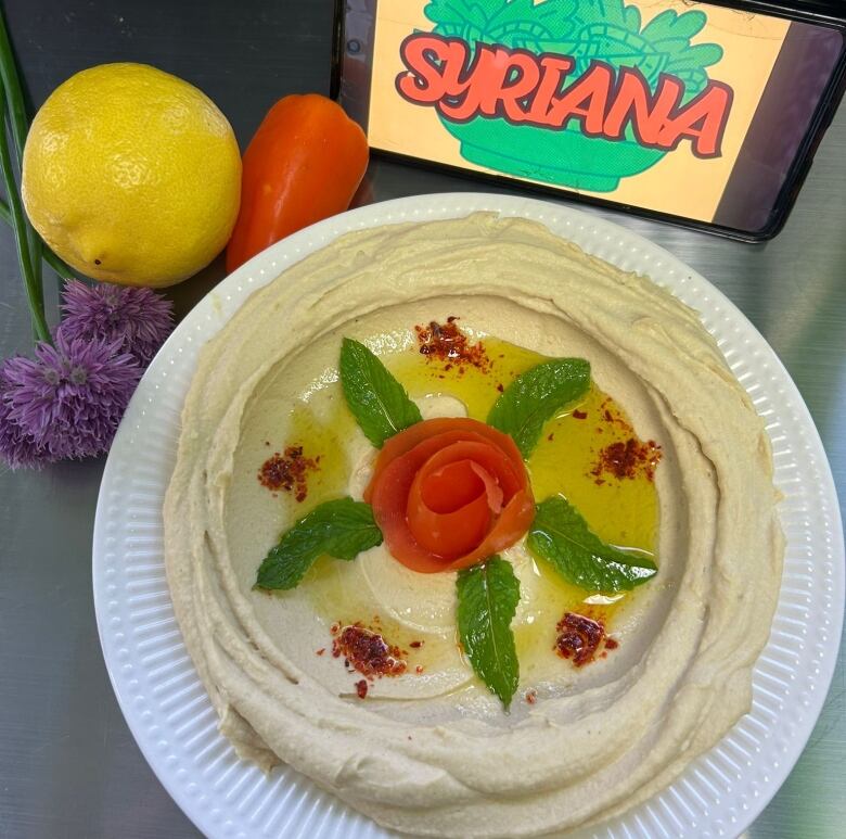 A plat of hummus with carrot in the center cut like a flower and mint leaves next to it. Next to the dish is a smartphone that reads 'Syriana'. 