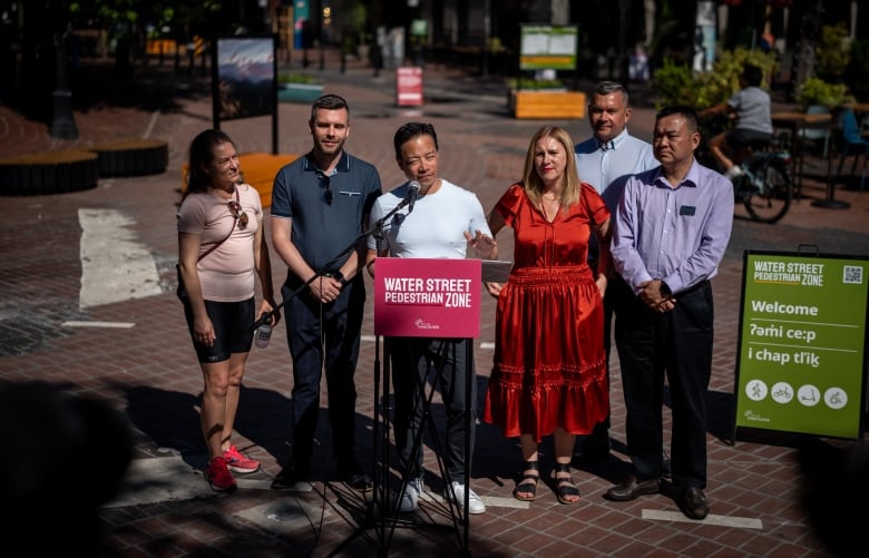 Six people stand behind a podium in a red brick square. 