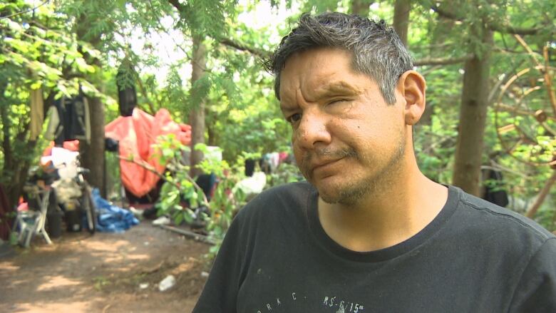 A man in a T-shirt stands in front of an encampment.