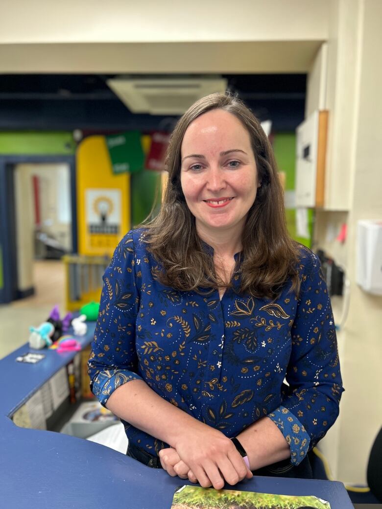 A young woman with long brown hair and wearing a blue and gold paisley shirt with the sleeves rolled up stands behind a blue counter in the Science East entryway and smiles at the camera for an upper body portrait.