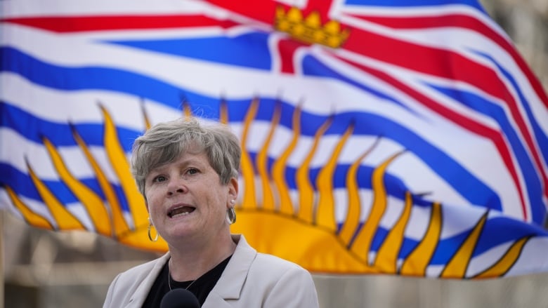 A woman stands at a microphone with a B.C. flag waving in the wind behind her. 