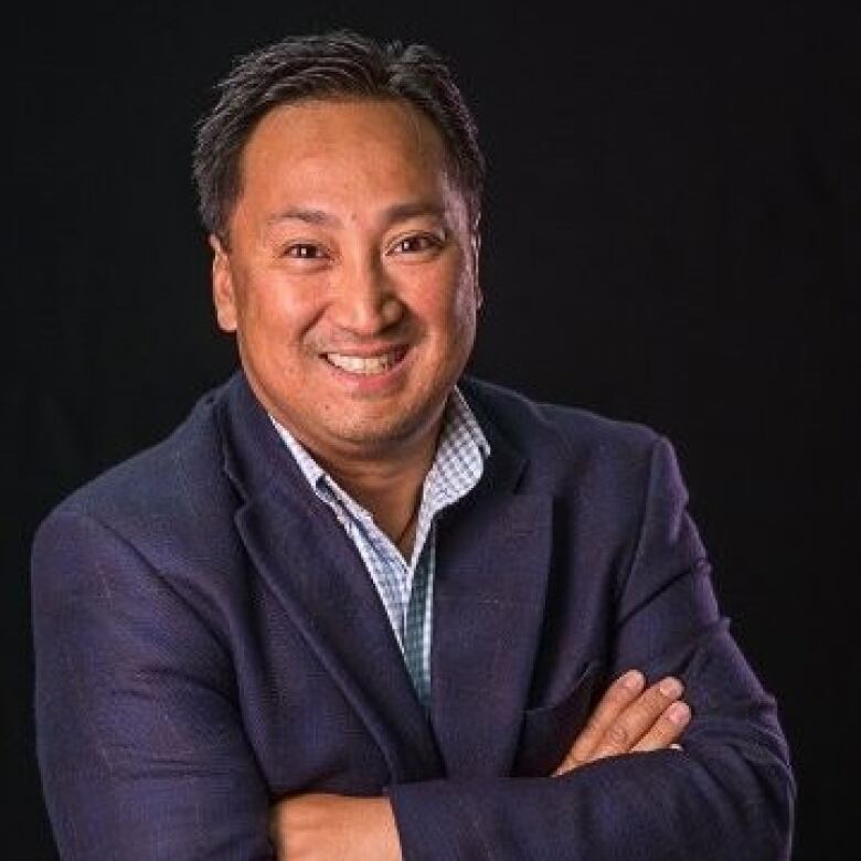 A smiling man with black hair wearing a blue suit and a blue and white checkered shirt crossing his arms