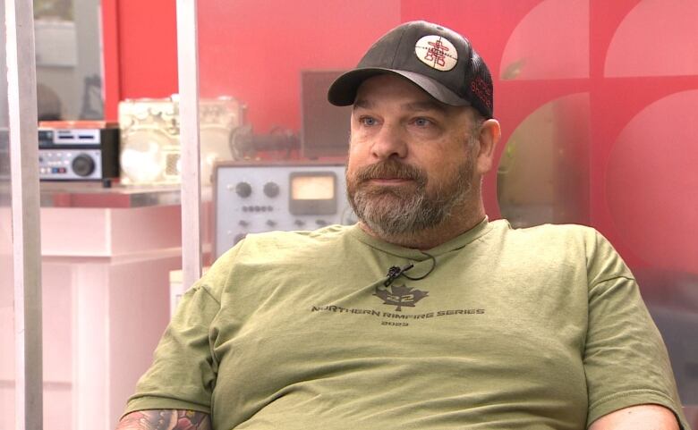 A sitting man with a greying brown beard wearing a green t-shirt and a black cap with crosshairs on it