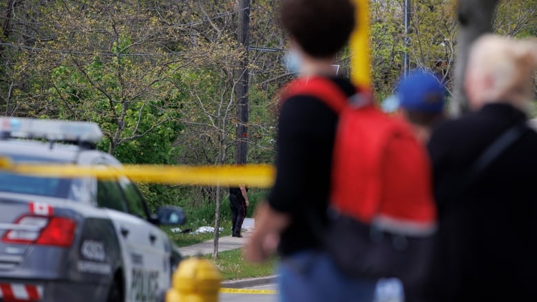 A boy with a red backpack is seen looking at a police car surrounded by yellow crime scene tape.