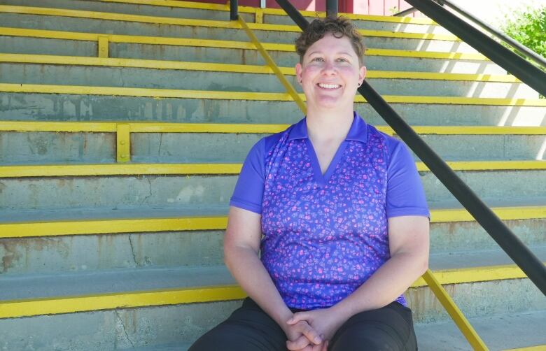 A woman with short hair and a purple top smiles while seated on a staircase.