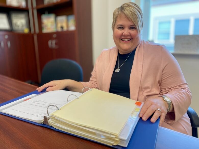 Raeanne Adams smiles at the camera, as she sits at her desk holding a binder. 