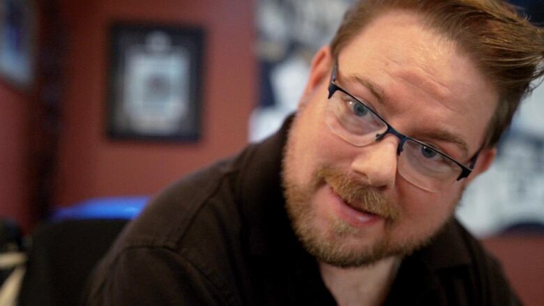 Close up of a man with short brown hair and glasses, wearing a black shirt, staring into the camera.