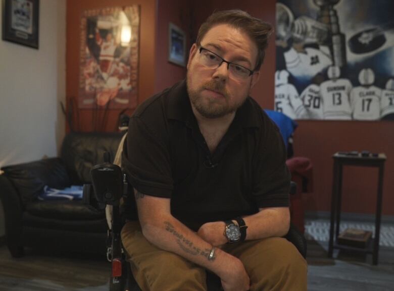 A man with short brown hair and glasses, wearing a black shirt and tan pants, sits in a wheelchair and looks into the camera.