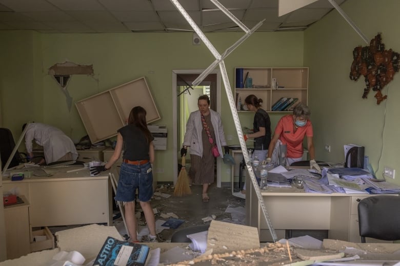 Four people are shown standing and walking inside what appears to be an office-like room, heavily damaged with debris on the ground and damage to the walls.