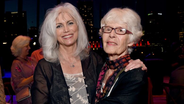 Two women with white hair smile and embrace side by side posing for a picture.