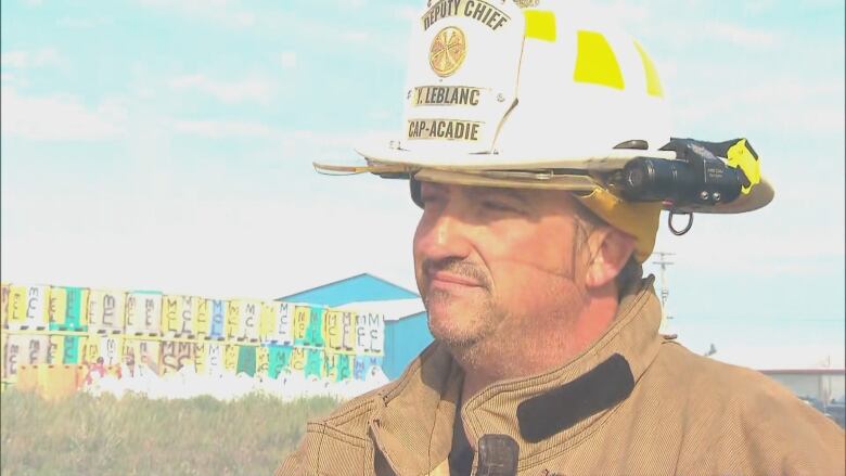 A man with a scruff of dark facial hair and wearing a beige coat and white firefighter had bearing the words LeBlanc and Cap-Acadie on the front of it looks forward at an angle to the left. In the background is a bright blue sky with some clouds and a stack of two rows of some kind of containers in bright yellow, white and green. You can also see a blue building with a white roof.