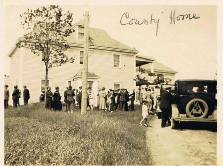 An old photo of a large two storey building. People stand outside. An old car is seen parked in the driveway by the home.