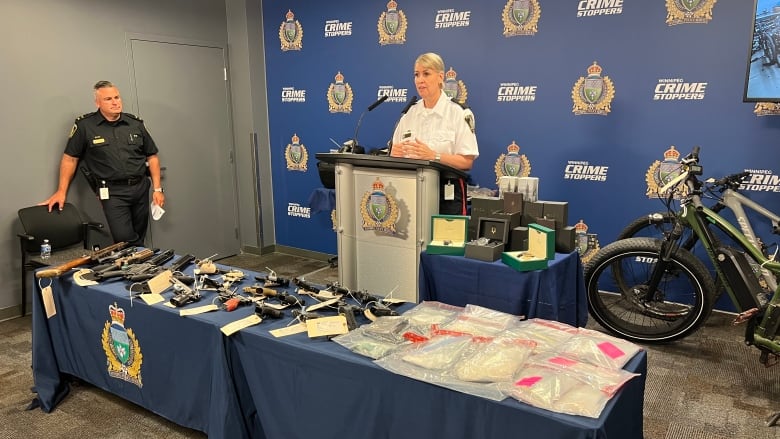 A woman speaks at a podium behind a table with various firearms and drug-related items on it.