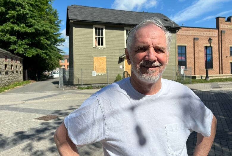 A smiling man wearing a white t-shirt stands in front of a boarded up building