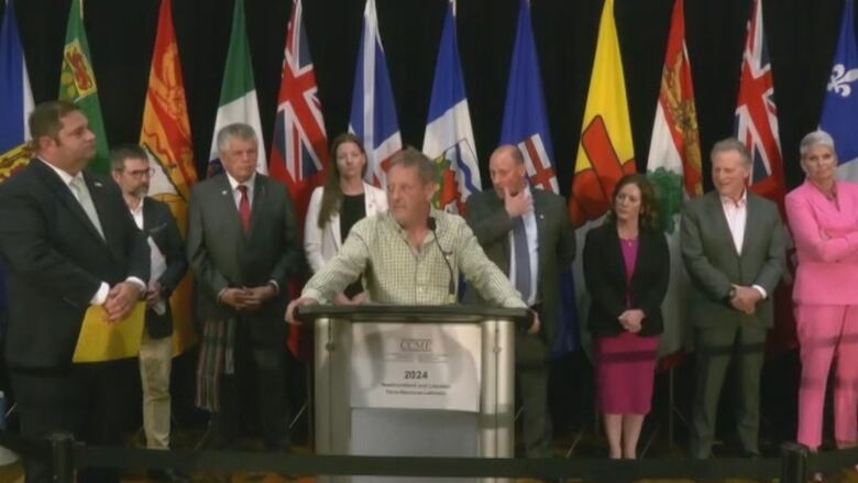 A man wearing a plaid shirt stands at a podium. Behind him, several government officials stare at him.