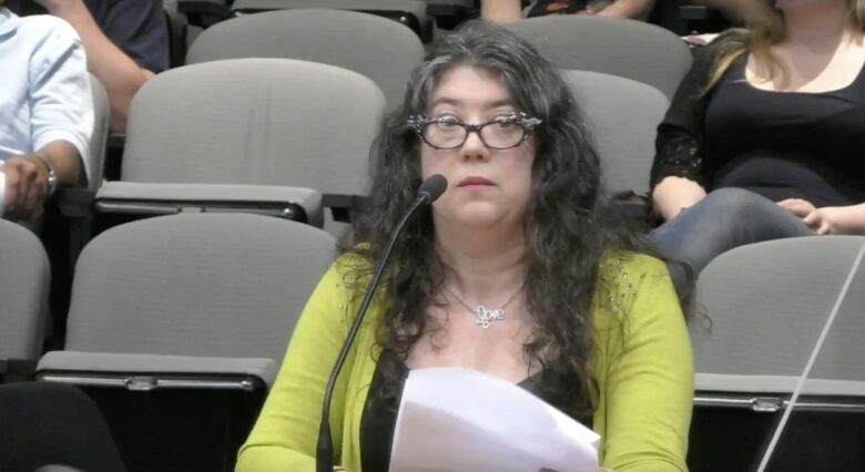 A white woman with black hair and black glasses speaks in council chambers.