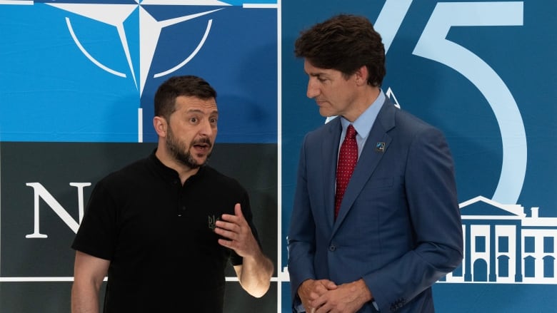 Two men stand in front of a blue backdrop; the one on the left in a black polo shirt and the one on the right in a blue suit with a red tie. 