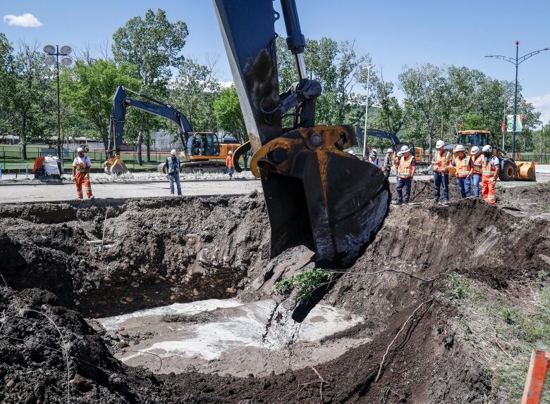 construction workers stand around a large hole filled with muddy water as a large piece of construction equipment digs into the hole.