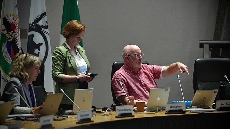 A woman stands behind a long desk checking her phone, while a woman beside her sits on a laptop and a man points off camera.