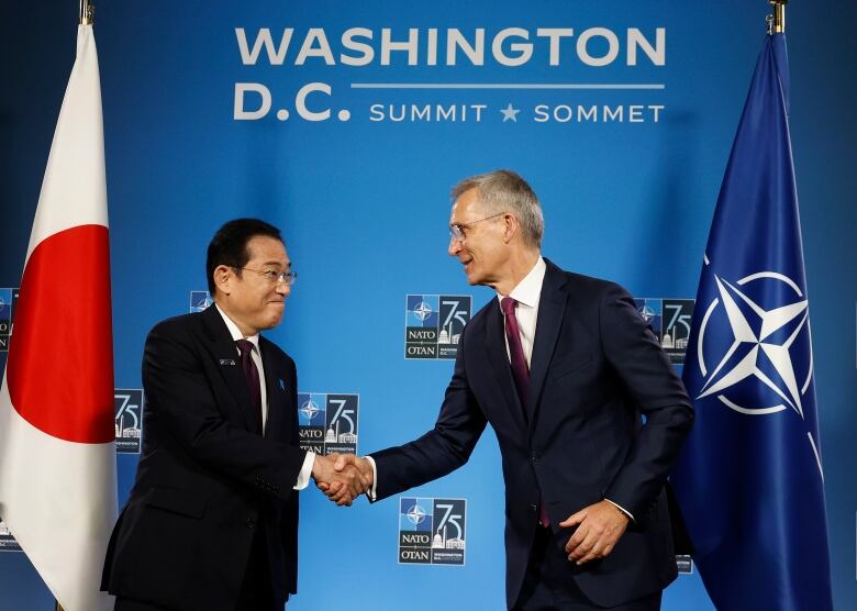 An Asiatic man and a Caucasian man in glasses shake hands on a stage in front of a number of flags.