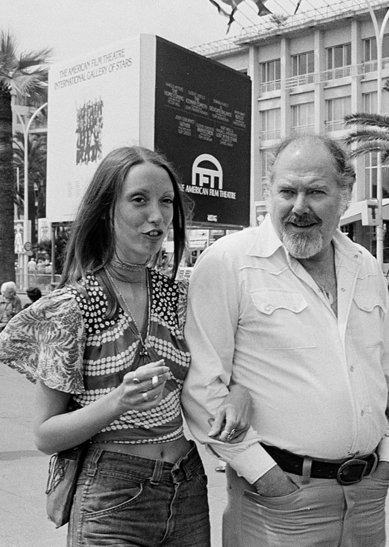 A thin woman is shown speaking while walking arm in arm with a balding, bearded middle-aged man in a black and white photograph.