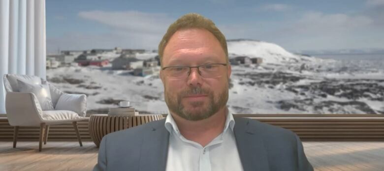 A man with brown hair and facial hair wearing glasses, a white shirt and blue suit sitting in front of a window overlooking an Arctic town