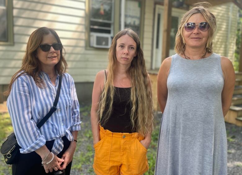 Three women stand outside in front of a house. Left wears a blue and white striped shirt, the one in the middle wears a black tank top and the one on the right wears a grey dress. 