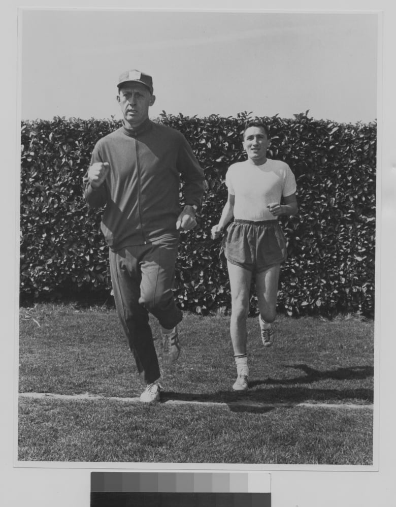 A man in a track suit and baseball cap demonstrates jogging technique to another man wearing shorts and a t-shirt in an archival photo from the 1960s.