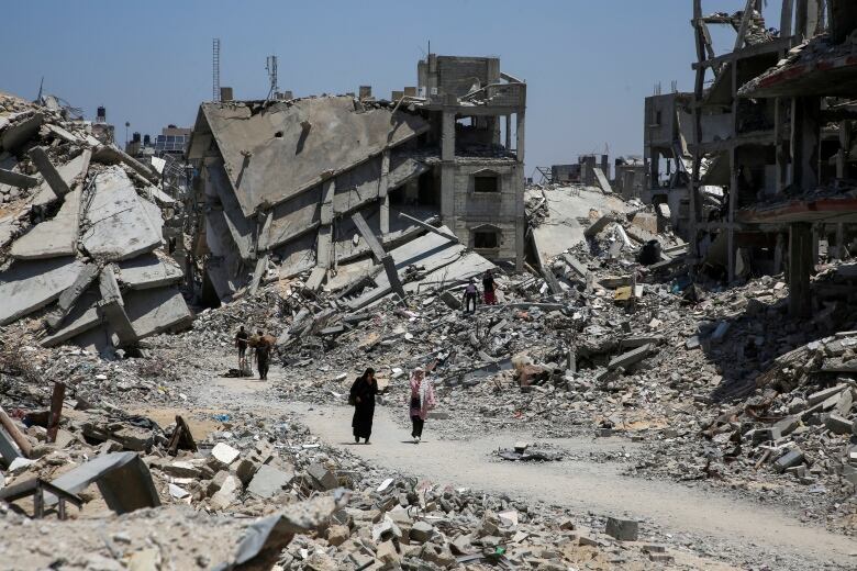 Two people are seen walking in the middle of a road with destroyed houses surrounding them.