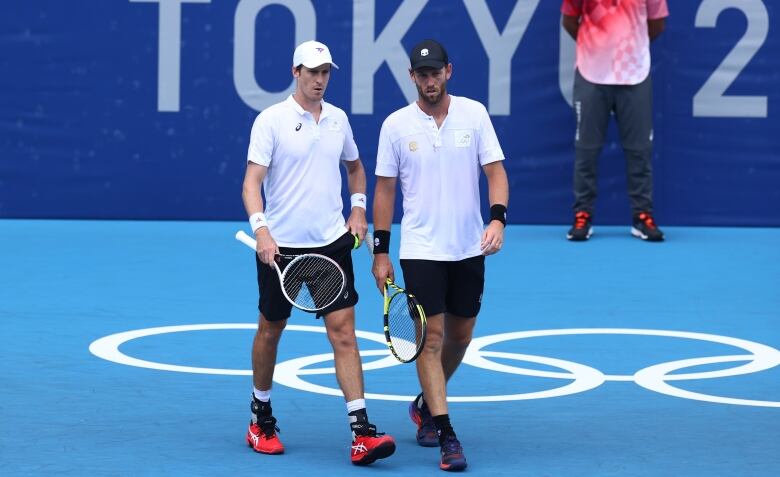 Two men with tennis rackets walk across a blue court. 