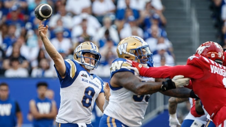 A man in a football uniform throws a football across the field while two other players tackle each other to his left. 