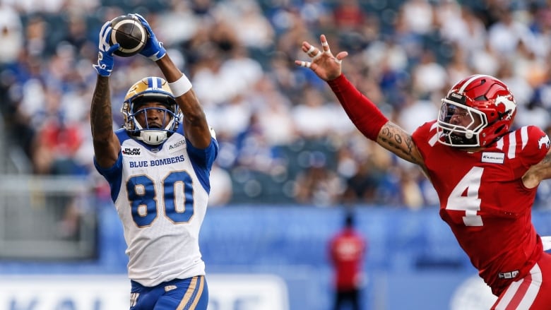 A man wearing a blue and white football uniform catches the ball as a player on the opposing team wearing red chases him. 