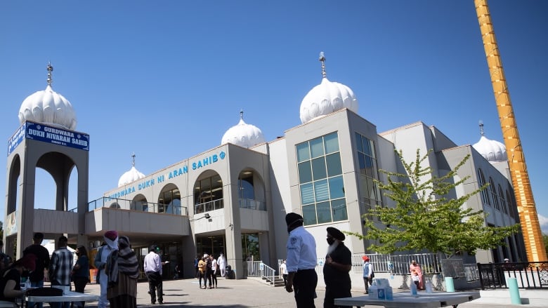 People gather outside a gurdwara, wearing facemasks, on a sunny day.