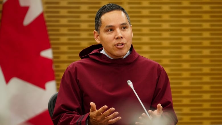 A man with black hair, wearing a reddish hooded shirt, speaks into a microphone beside a Canadian flag.